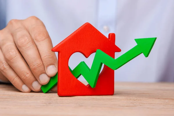 Αυξανόμενες τιμές κατοικίας. Ο άνθρωπος είναι κρατώντας το πράσινο βέλος επάνω στο χέρι και σπίτι στο τραπέζι — Φωτογραφία Αρχείου