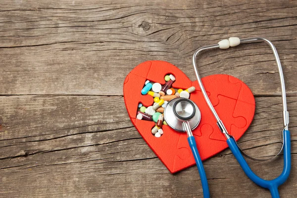 Cuore puzzle rosso e pillole di colore e stetoscopio su uno sfondo di legno. Concetto trattamento di pillole per malattie cardiache — Foto Stock