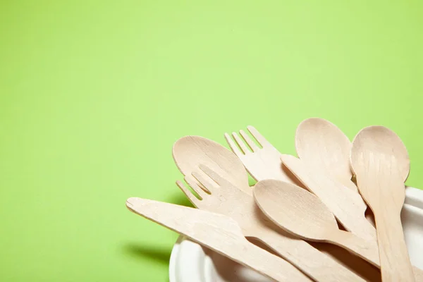 Eco-friendly disposable utensils made of bamboo wood and paper on a green background. Draped spoons, fork, knives, bamboo bowls