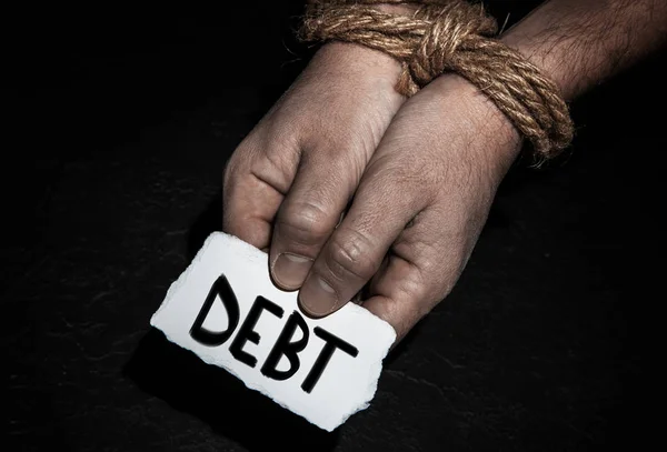 Inscripción de la deuda en papel en un hombre con las manos atadas con cuerda sobre fondo negro — Foto de Stock