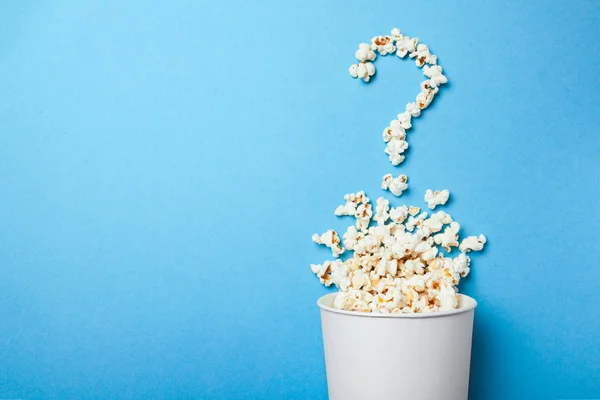 Popcorn wird in Form eines Fragezeichens aus einem weißen Pappbecher auf blauem Hintergrund ausgeschüttet. Wie wählt man einen guten Film zum Anschauen aus? — Stockfoto