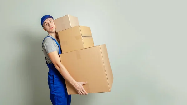 Day moving. Delivery of goods from shopping in the online store. Loader or courier carries heavy cardboard boxes against gray wall