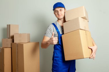 Hareketli bir gün. Online mağazada alışveriş teslim mal. Yükleyici veya kurye gülümsüyor ve büyük başparmak ortaya çıkıyor ve gri duvara karton kutular taşır