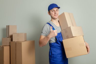 Hareketli bir gün. Online mağazada alışveriş teslim mal. Yükleyici veya kurye gülümsüyor ve büyük başparmak ortaya çıkıyor ve gri duvara karton kutular taşır