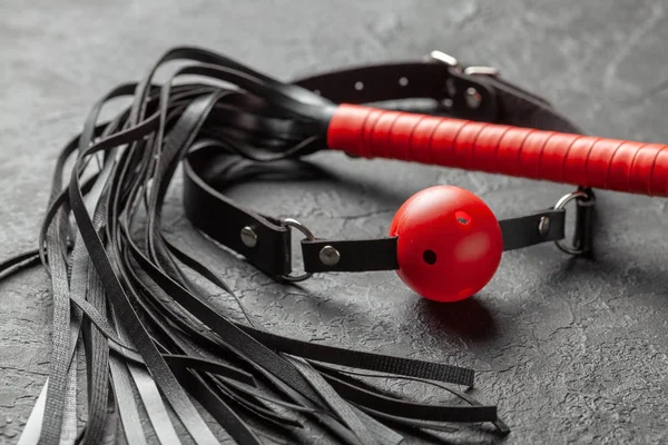 BDSM juguetes sexuales para adultos. Mordaza con bola roja y látigo sobre un fondo negro . — Foto de Stock