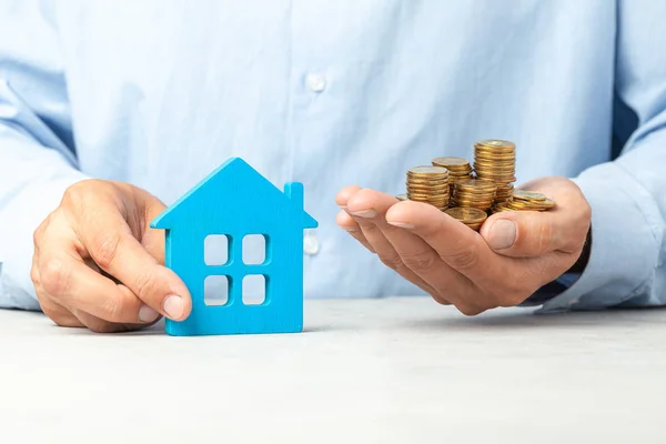 El hombre de camisa azul sostiene una casa y un montón de monedas. El concepto de comprar casa, impuestos sobre bienes raíces, seguro de vivienda — Foto de Stock