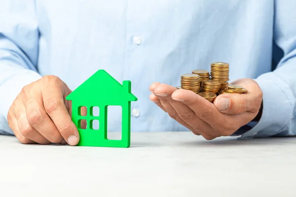 El hombre de camisa azul sostiene una casa y un montón de monedas. El concepto de comprar casa, impuestos sobre bienes raíces, seguro de vivienda — Foto de Stock