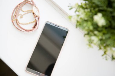 Akıllı telefon ve kadın mücevherleri yeşil bir çiçeğe karşı beyaz bir pencere eşiğinde yatıyor.