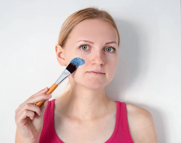 Vacker blond kvinna med blå lera ansiktsmask tillämpas av kosmetolog. flicka med en mask på en kind ler — Stockfoto