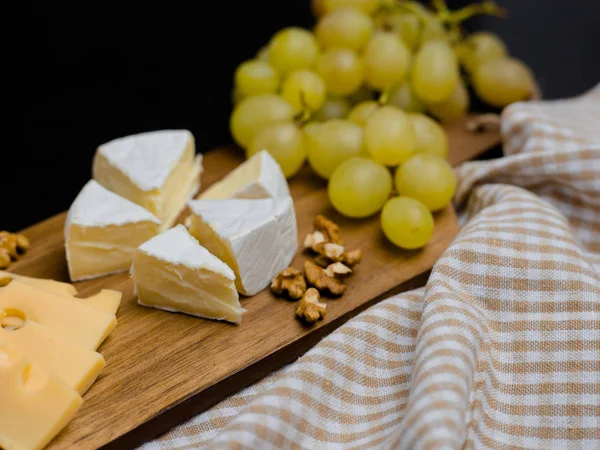 Rebanada de queso, nueces y uvas en tabla de cortar de madera. Queso Camembert y queso edam con tela a cuadros. Comida para el vino y romántico — Foto de Stock