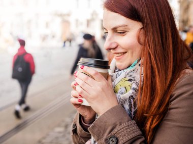Kahve fincanı tutan ve bulanık sokakta gülümseyen bir kadın. Güneşli bir bahar öğleden sonra. Kırmızı manikür.