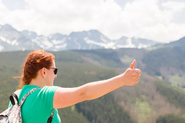 Junge Frau mit Rucksack, die Daumen hoch gibt. Wander- und Tourismuskonzept. Kopierraum. eine junge hübsche rothaarige Frau, die auf einem Hintergrund von Bergen steht — Stockfoto