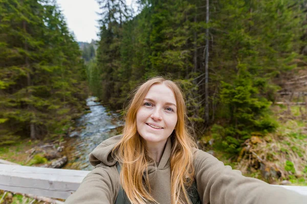 Güzel genç bayan yürüyüşçü dağ ormanında selfie çekiyor. Kameraya bakıp gülümse. Seyahat ve aktif yaşam konsepti. Açık havada. — Stok fotoğraf