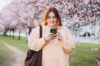 Baharın güzel bir gününde telefonunu kullanan genç bir kadın çiçek açan bir ağacın ortasında dikiliyor.