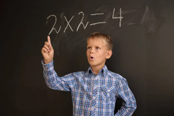 European boy student thinks on the solution of mathematical problems, standing at a black school Board