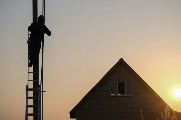Eletricista realiza instalação de equipamentos elétricos no suporte da linha de energia perto de uma casa de madeira à noite ao pôr do sol — Fotografia de Stock