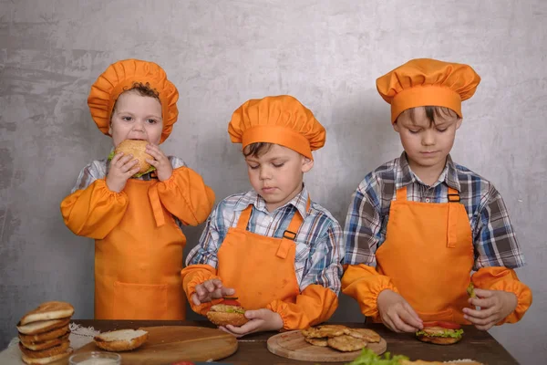 Tres chicos lindos en trajes cocineros dedicados a cocinar hamburguesas caseras. tres hermanos preparando la cena familiar — Foto de Stock
