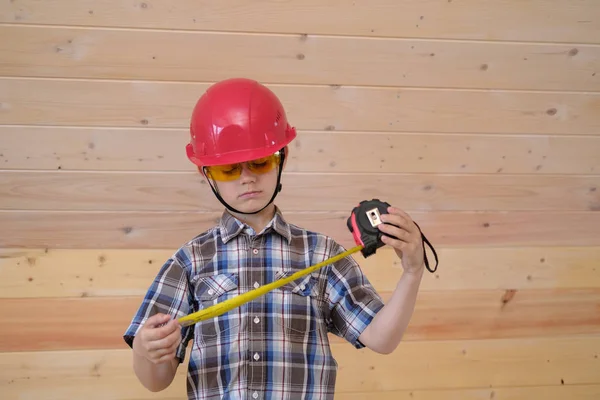 Un niño lindo en un casco protector y gafas mide la pared de una nueva casa de madera con una cinta métrica de construcción —  Fotos de Stock