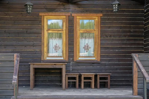 Drewniana brązowa fasada zabytkowego domu z dwoma oknami. drewniany stół i stołki na wal — Zdjęcie stockowe