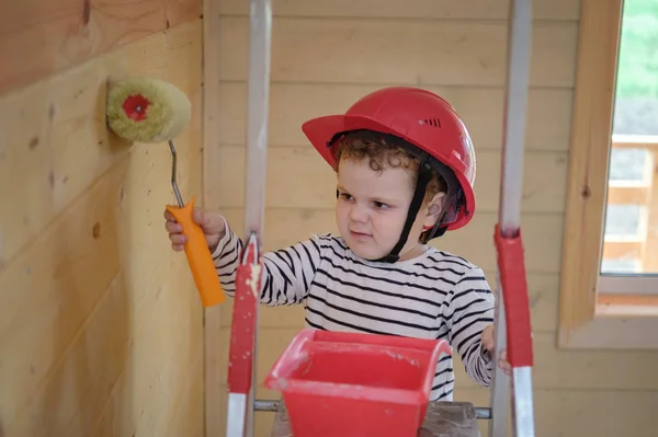 Милый маленький мальчик в защитном шлеме красил стену деревянного дома роликом, стоящим на стремянке — стоковое фото
