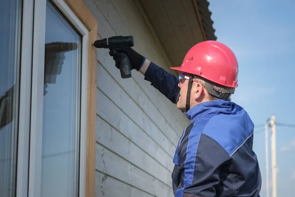 Europese mannelijke bouwer in overalls assembleert een raam behuizing van een houten huis met een schroevendraaier. bouw en reparatie van houten frame huizen — Stockfoto