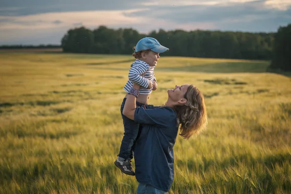 Happy mom throws her son up in her arms on a barley field on a summer evening — Stock Photo, Image