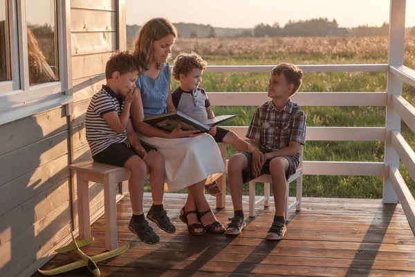 Mor med tre barn läser en bok på sommarterrassen i ett hus på landet. familjebok läsning — Stockfoto