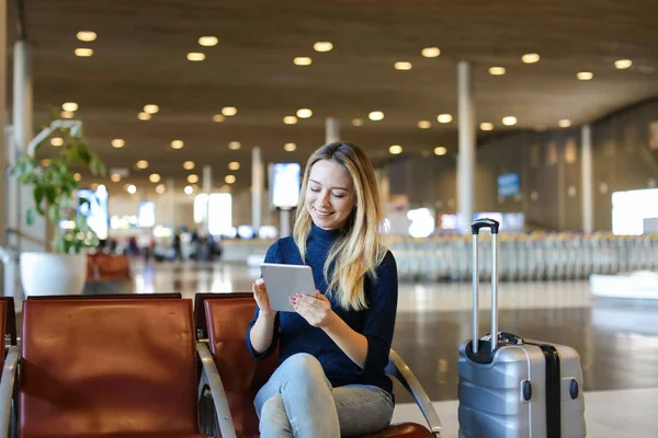 Female person sitting in airport waiting room with valise and using tablet.