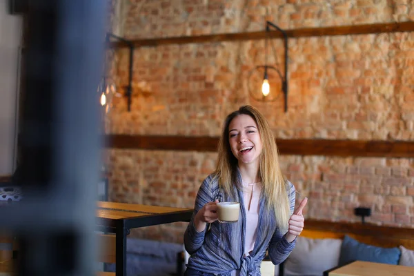 Zufriedene Mädchen am Tisch mit einer Tasse Cappuccino in einem gemütlichen Café mit ungewöhnlichem Interieur. — Stockfoto