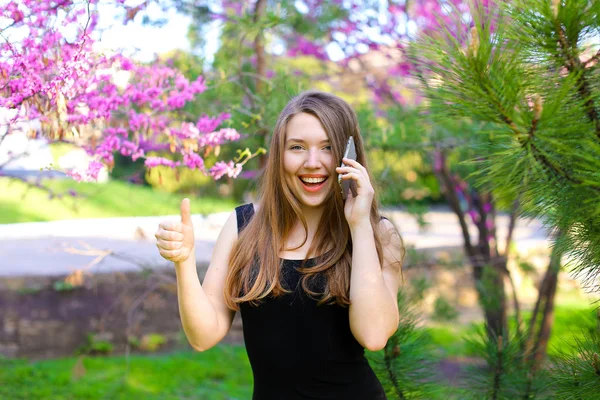 Hermosa mujer hablando por teléfono inteligente y mostrando pulgares hacia arriba en el parque de primavera . — Foto de Stock