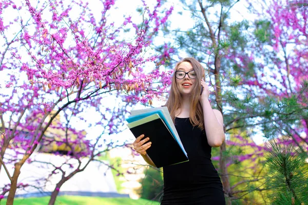 Young female secretary in glasses talking by smartphone in blooming park with documents.