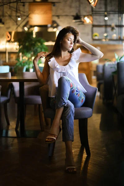 Стильная женщина сидит в кафе с поднятой рукой и в белой блузке с этническими джинсами . — стоковое фото