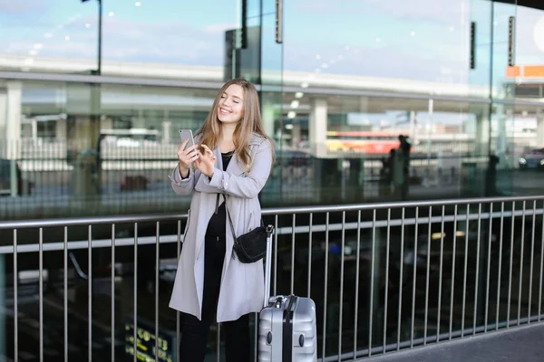 European woman chatting and browsing by smartphone near valise and airport.
