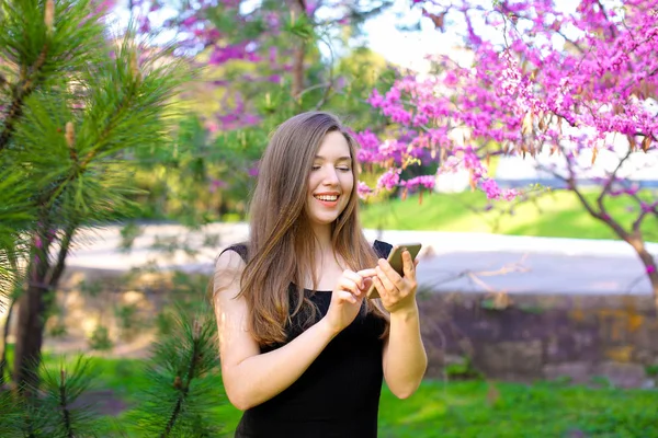 Cheerful girl using smartphone in garden with blossom background. — Stock Photo, Image