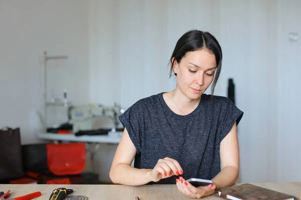 Ευρωπαϊκή craftswoman κάθεται στο ατελιέ και χρησιμοποιώντας το smartphone, χειροποίητα δερμάτινα είδη. — Φωτογραφία Αρχείου