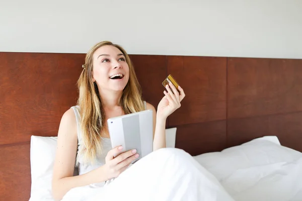 Fiatal, boldog asszony lesz a tábla- és betéti kártya online vásárlás, ült az ágyban. — Stock Fotó