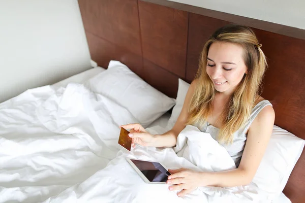 Young european woman booking tickets online by tablet and gold debit card, sitting in bed.