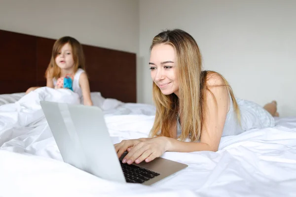 Joven madre acostada en la cama con su hijita y usando laptop . — Foto de Stock