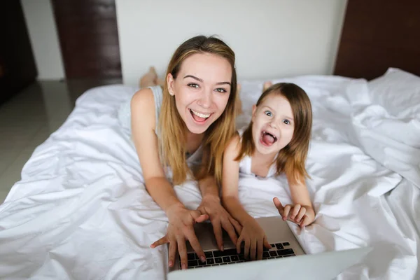 Feliz caucásico persona femenina utilizando el ordenador portátil con su hija pequeña y acostado en la cama . — Foto de Stock