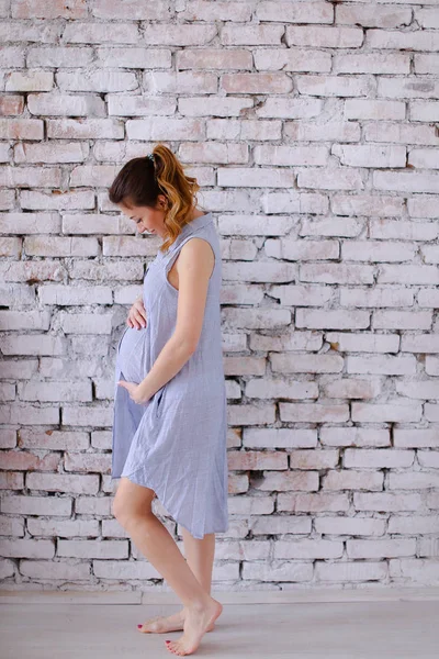 Tuğla duvar arka planda duran ve göbek tutan mavi elbise giyen hamile cauca Avrupa sian kadın. — Stok fotoğraf