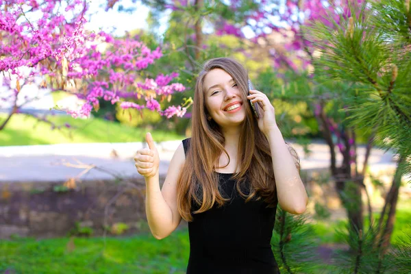 Junge Frau spricht mit Smartphone und zeigt Daumen hoch im Frühlingspark. — Stockfoto