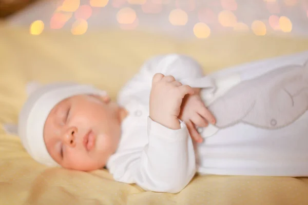 Kleines nettes Baby in weißer Mütze auf Decke liegend, gelbes grelles Licht im Hintergrund. — Stockfoto