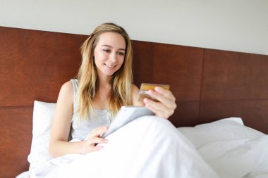 Tablet ve bankamatik kartı ile alışveriş online, yatakta oturan genç kadın.