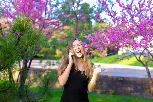 Ładna kobieta mówiąc przez smartfon w parku w pobliżu kwitnących drzew. — Zdjęcie stockowe