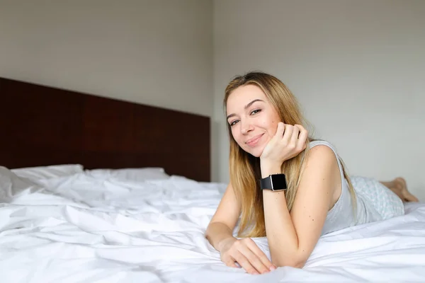 年轻的白种女人使用 smartwatch 和躺在床上与白色亚麻布在早上. — 图库照片