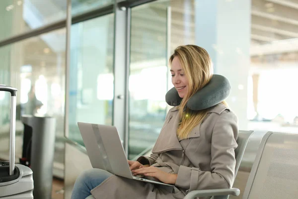 Female person sitting in waiting room with laptop near valise, using neck pillow.