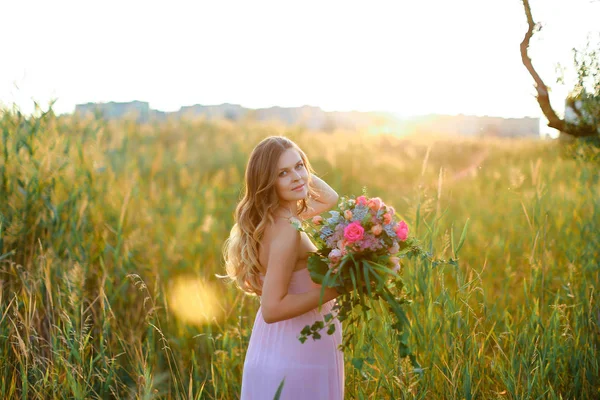 Pregnant young caucasian woman wearing pink dress with bouquet of flowers standing in steppe background. — Stock Photo, Image