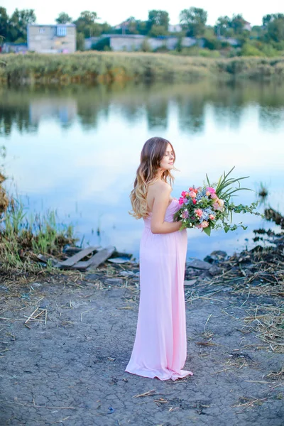 Pregnant blonde woman wearing pink dress with bouquet of flowers standing near lake. — Stock Photo, Image