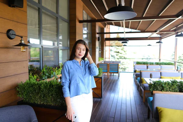 Kvinna står vid café med smartphone och bär jeans skjorta och vita byxor. — Stockfoto