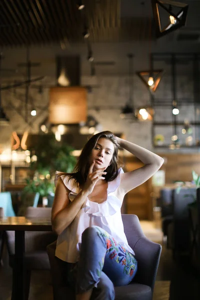 세련 된 여 자가 제기 손으로 카페에 앉아 민족 청바지와 흰색 블라우스를 입고. — 스톡 사진